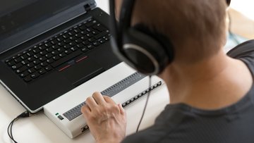 Ein Mann mit Headset sitzt vor einem Laptop und tätigt Eingaben über eine Braille-Tastatur.