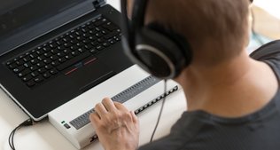 Ein Mann mit Headset sitzt vor einem Laptop und tätigt Eingaben über eine Braille-Tastatur.