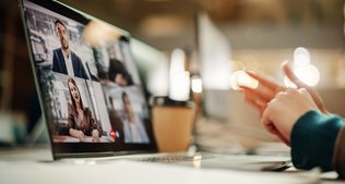 Ein Laptop auf einem Tisch zeigt eine Videokonferenz und die Hände der vor dem Laptop sitzenden Person.