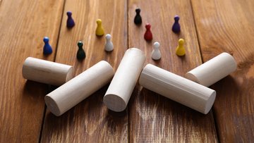 Bunte Spielfiguren stehen auf einer Holzfläche, gegenüber befinden sich große, zylinderförmige Bausteine.