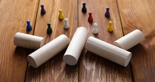 Bunte Spielfiguren stehen auf einer Holzfläche, gegenüber befinden sich große, zylinderförmige Bausteine.